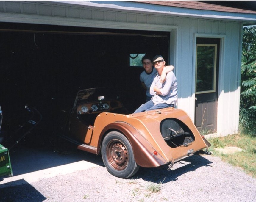 Two people next to a vintage car parked in a garage.