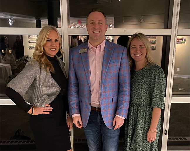 Three smiling people in business attire standing in front of a glass window.