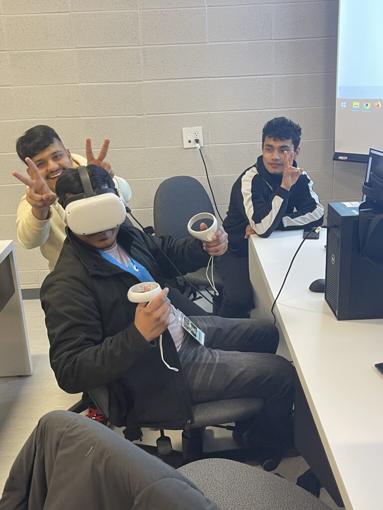 Three males smiling and sitting in front of computers in a classroom. One of them is wearing vr goggles