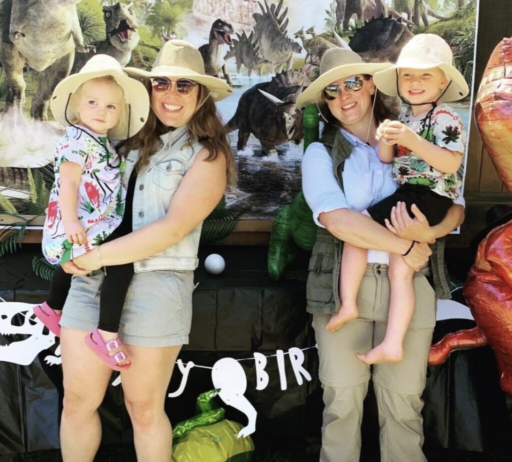 Two adults each hold a child in their arms while standing in front of a dinosaur backdrop. 