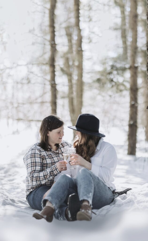 Two people sit on the ground in a snowy forest and cheers champagne. 