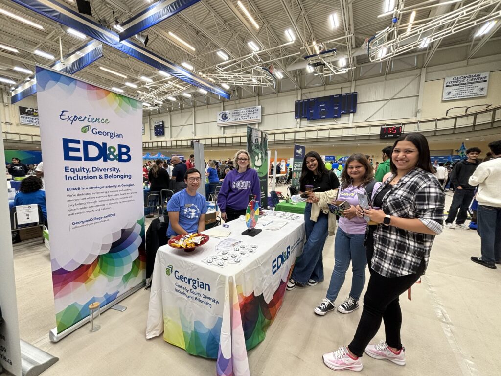 Five people gather around a table and a poster reading "Experience Georgian. EDI&B Equity, Diversity, Inclusion & Belonging" at a vendor event inside a gym. 