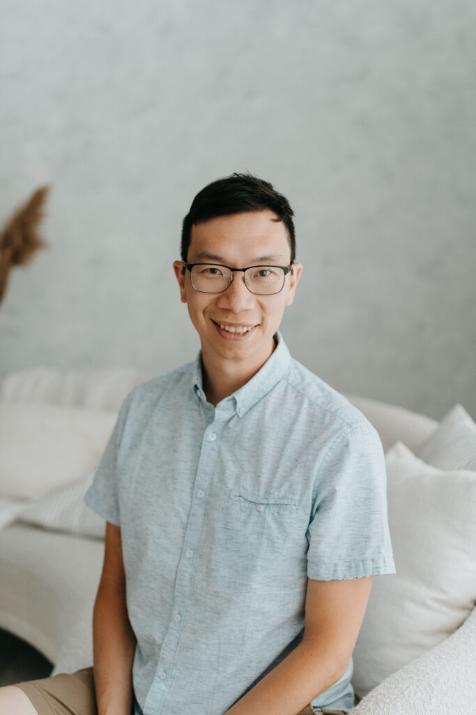 A person with short black hair, glasses, blue shirt and tan shorts, sits on a white couch and smiles. 