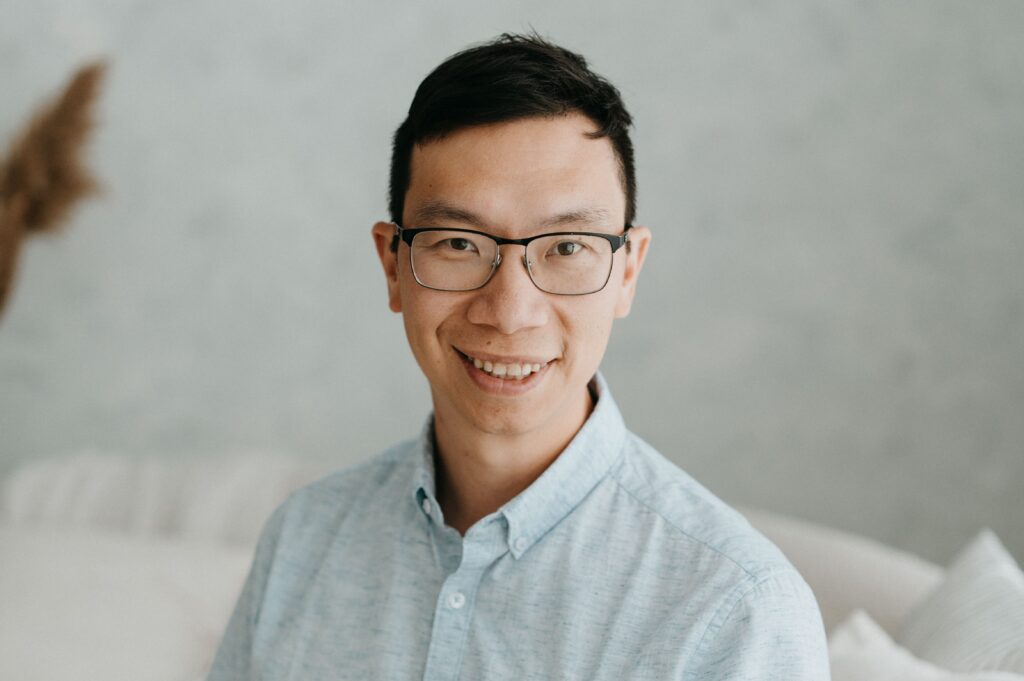 A person with short black hair, glasses and blue shirt, smiles.