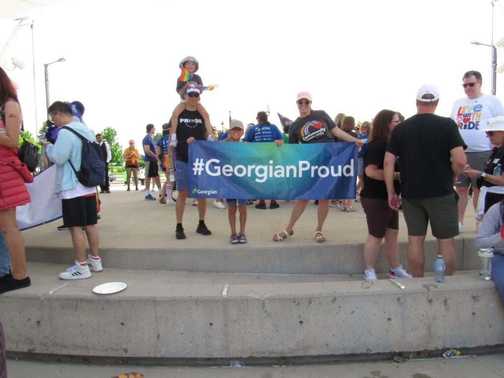 Two adults and two children stand on a platform and hold a sign reading "#GeorgianProud" surrounded by a crowd of people. 