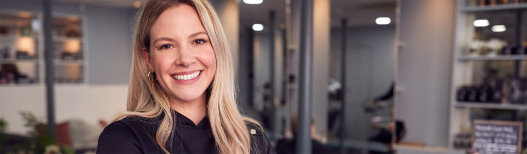 An esthetics and spa manager wearing a black scrub shirt, with long, blonde hair and arms folded, smiling while standing in a salon