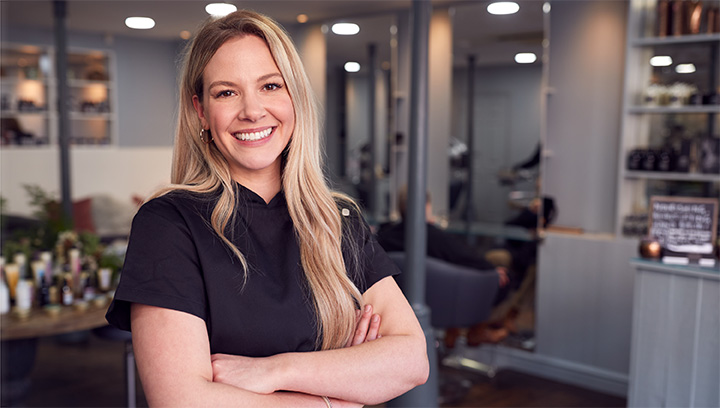 An esthetics and spa manager wearing a black scrub shirt, with long, blonde hair and arms folded, smiling while standing in a salon