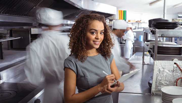 An event manager with brown, curly hair in a heather gray dress taking notes on a clipboard inside a commercial kitchen
