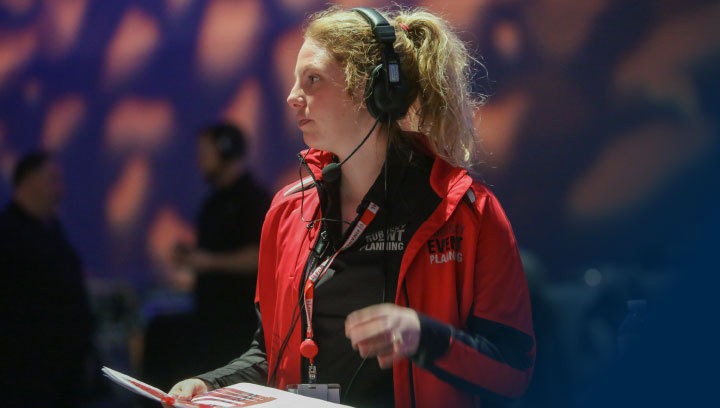An event manager wearing a branded shirt, jacket and lanyard with headphones and a clipboard onsite at an event