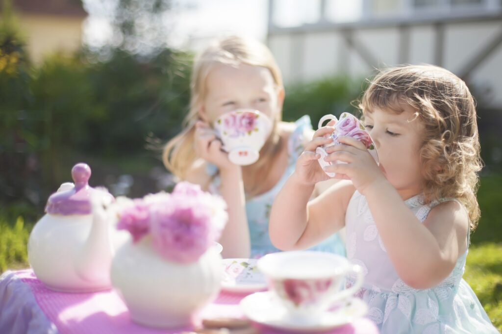 Two children in dresses sit outside at a table covered in a teapot and cups and they sip from teacups.