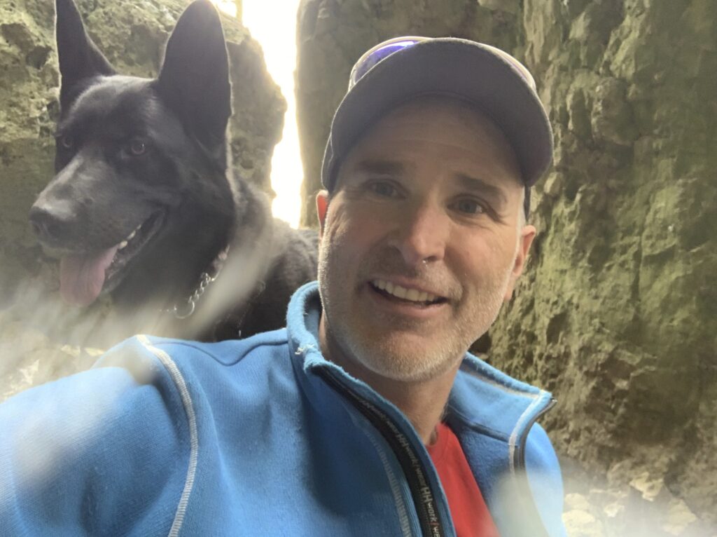 An adult takes a selfie with a dog in front of some large rocks.