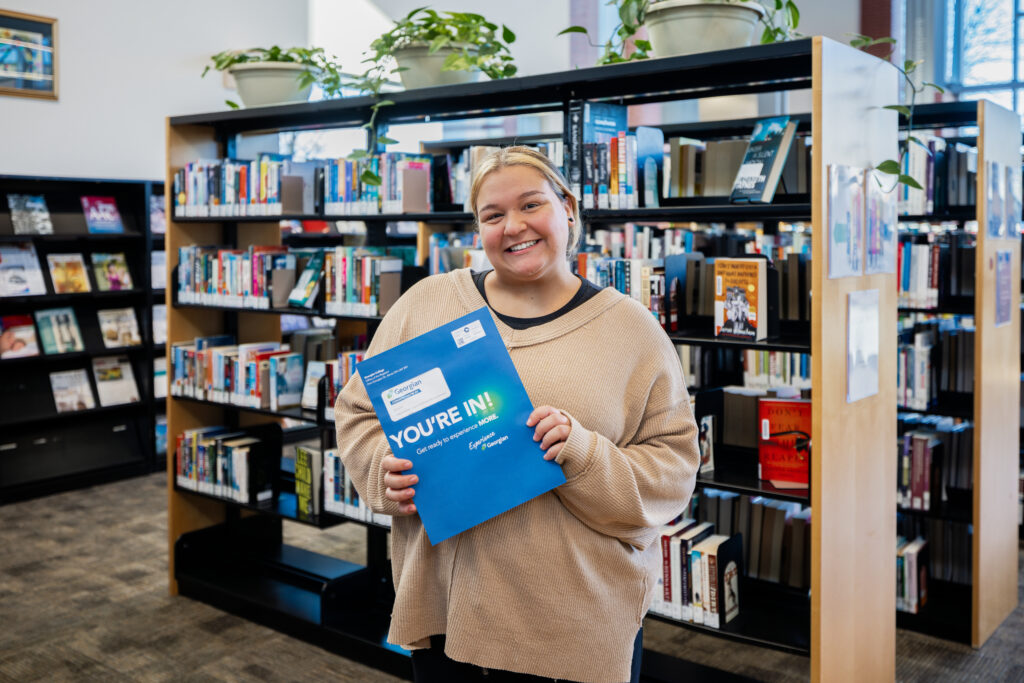 A person holds a blue envelope reading "You're in!" while standing in a library. 