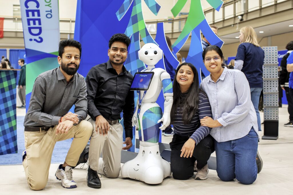 Four people kneel down on a floor next to a robot and smile. 