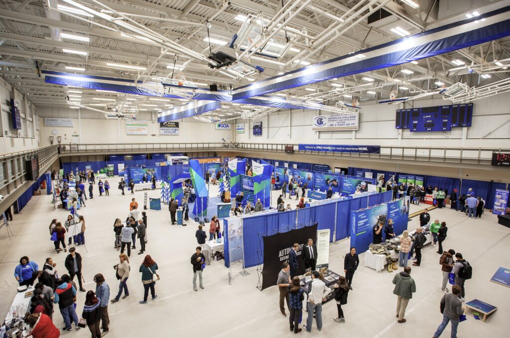 An aerial view of a gymnasium with blue tables and signage set up throughout and people walking around the room. 