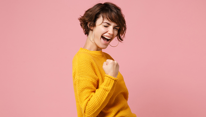 Person with short, brown hair in a yellow knit sweater with eyes closed, smiling with fist clenched in a success pose