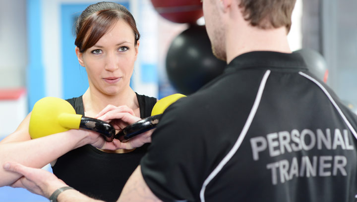 A personal trainer in a golf shirt stabilizing a client's arms while holding two yellow kettle bell weights in a fitness centre