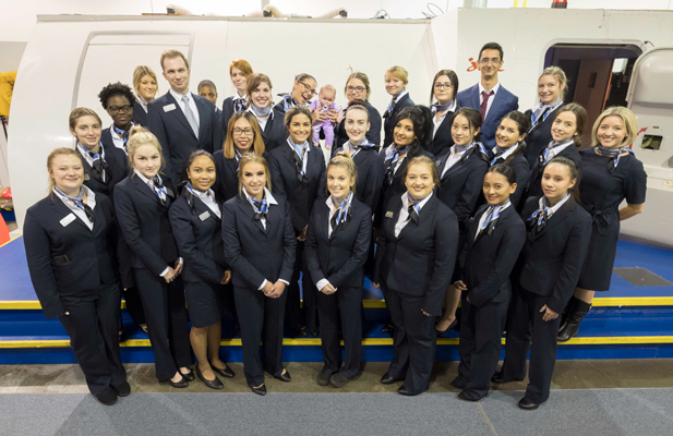 A group of Flight Services students in flight attendant uniforms standing on a platform in front of a Jazz Aviation airplane simulator