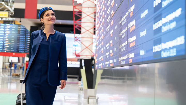 A flight attendant in a navy blue dress, blazer and hat carrying a suitcase behind them and looking up at the flight schedule screen in an airport