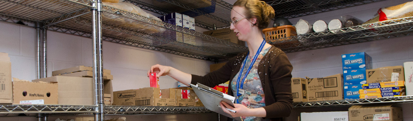 A Food and Nutrition Management students wearing a hair net while holding a carton of soup, pen and checklist in a pantry