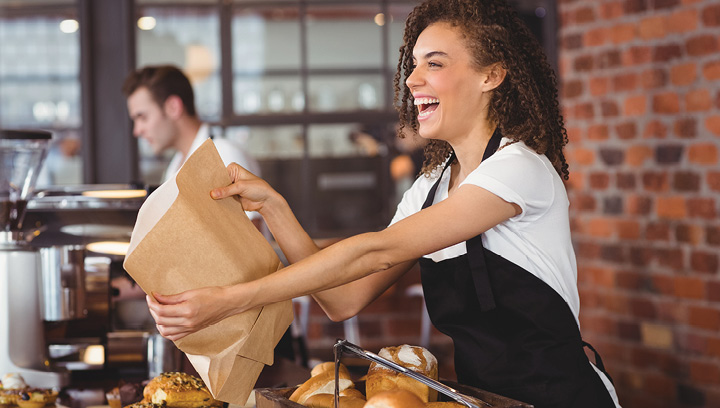 A food service worker with brown curly hair, in a white short-sleeved t-shirt and a black apron, smiling and holding out a paper bag in a cafe/bakery setting