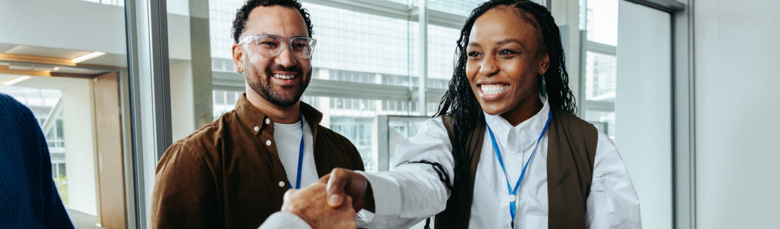 Two individuals with event badges stand in a modern indoor space while one shakes hands with another person, representing professional networking and relationship building.