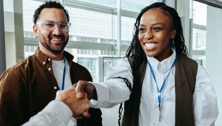 Two individuals with event badges stand in a modern indoor space while one shakes hands with another person, representing professional networking and relationship building.
