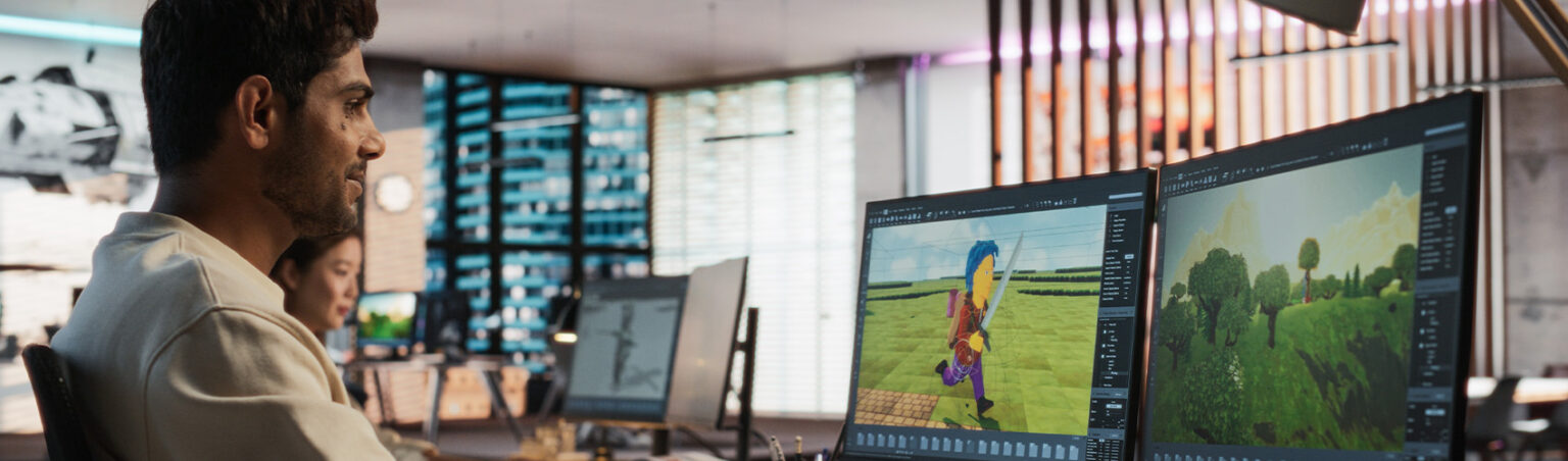 A man in an office works at a dual-monitor setup, designing a 3D video game character and environment. One screen shows a character holding a sword, while the other displays a landscape with trees and mountains.