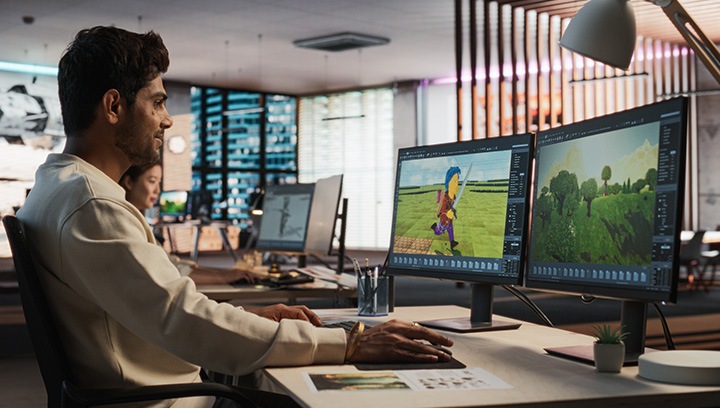 A man in an office works at a dual-monitor setup, designing a 3D video game character and environment. One screen shows a character holding a sword, while the other displays a landscape with trees and mountains.
