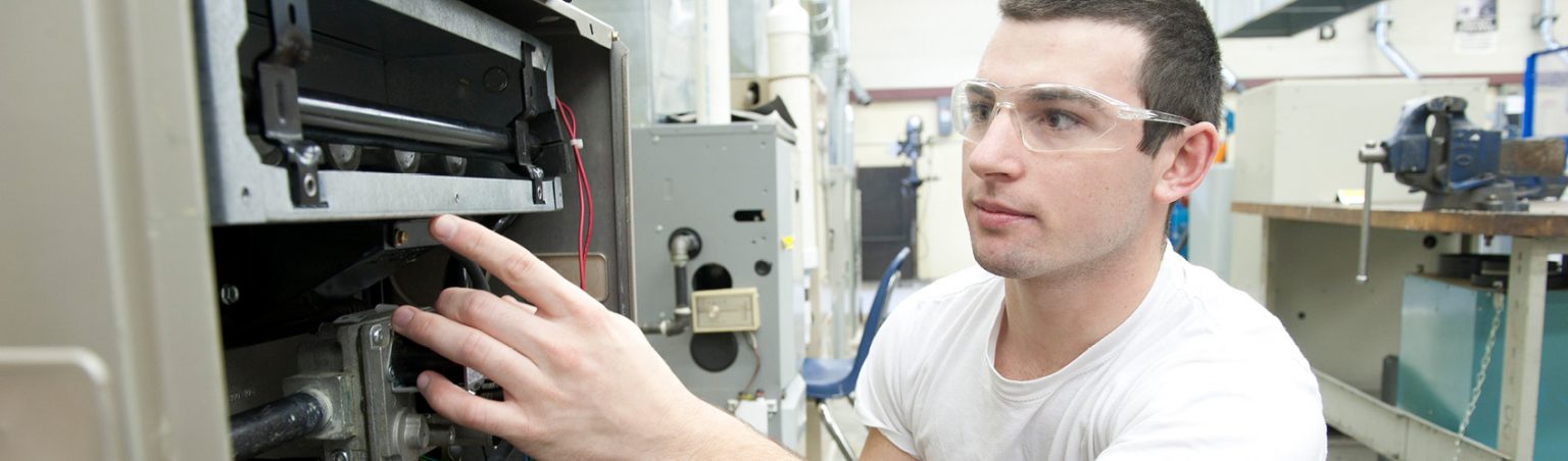 A Gas Technician student wearing jeans, a white t-shirt and safety glasses repairing a piece of equipment in a lab