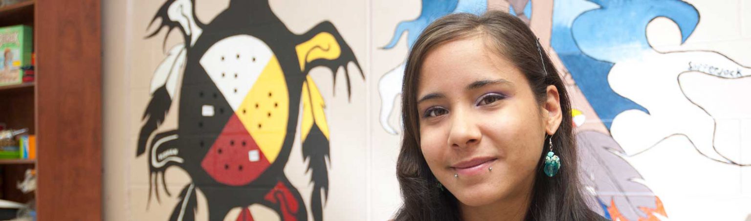 Indigenous Studies student at СŶƵ standing in front of a mural featuring an Indigenous medicine wheel