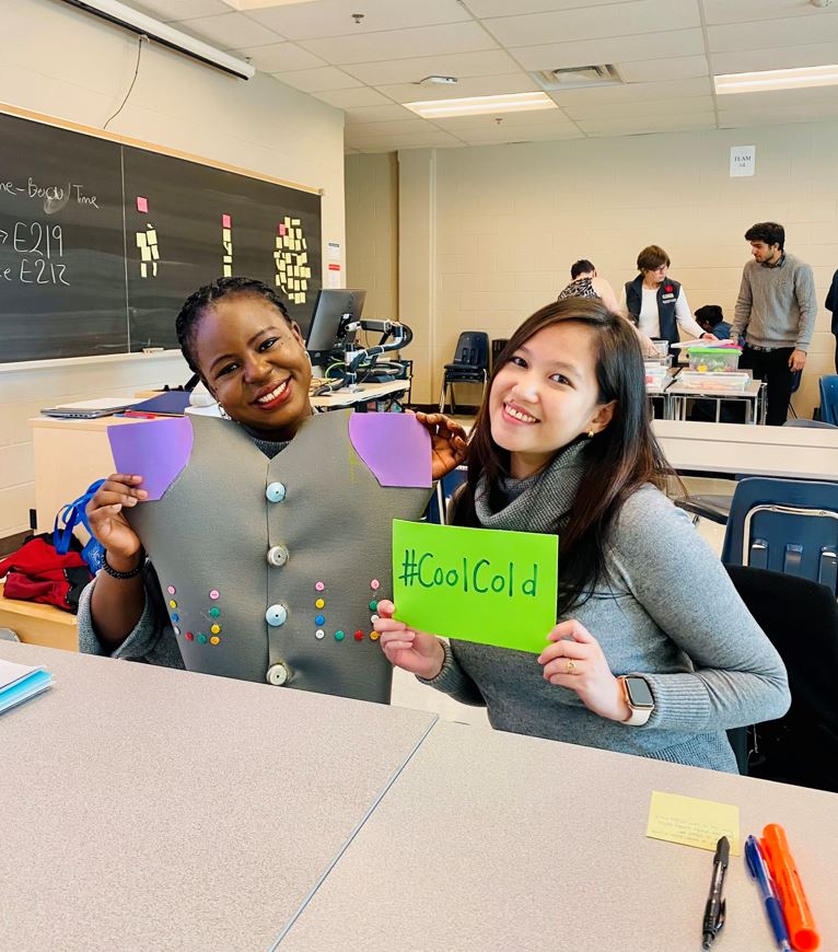 Two people sitting in a classroom at a desk hold up paper designs in front of them. One reads #CoolCold.