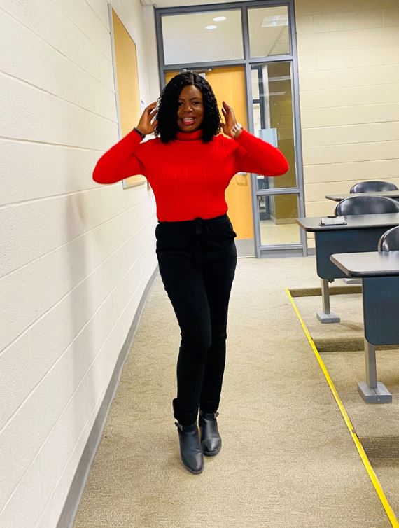 A person wearing a red sweater, black pants and black boots, poses with their hands up by their head while standing in a classroom.