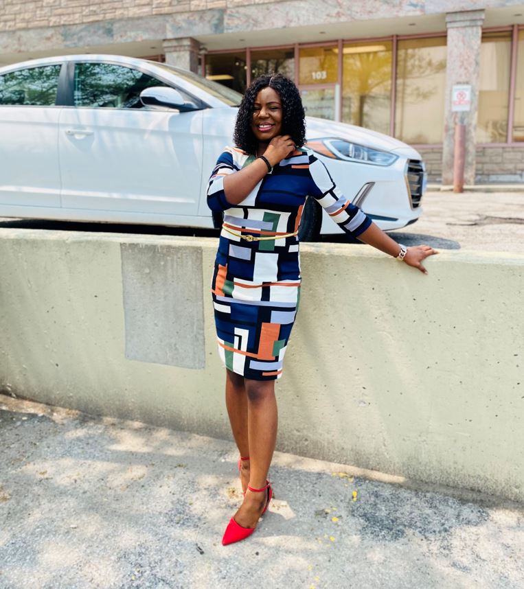 A person wearing a dress and red shoes smiles and stands outside next to a road.