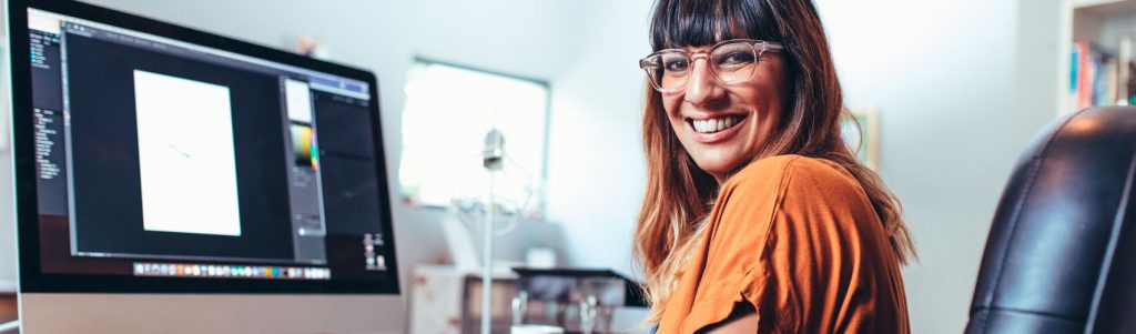 Graphic Design student with brown hair, glasses, an orange shirt and forearm tattoos using Adobe Photoshop on a Mac computer