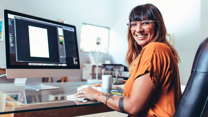 Graphic Design student with brown hair, glasses, an orange shirt and forearm tattoos using Adobe Photoshop on a Mac computer