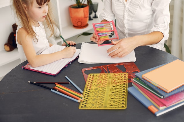 child and adult sitting at a round wooden table with notebooks, coloured pencils and stencils