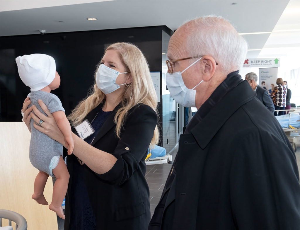A woman with long blonde hair holding up a baby mannequin while an older man stands beside her.