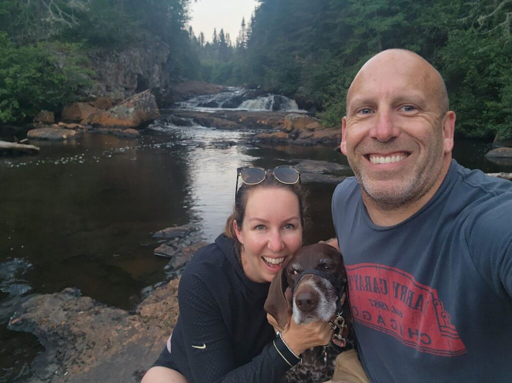 Two people and a dog stand together for a selfie in front of a river in the woods.