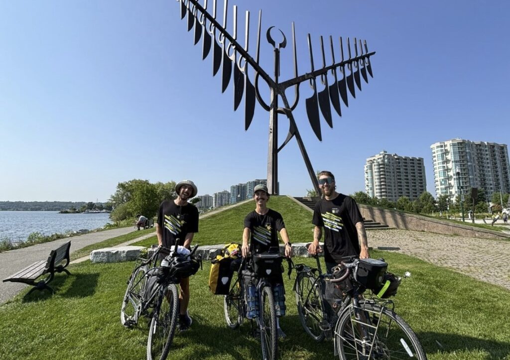 Three people with bikes stand in front of a large sculpture along a waterfront.