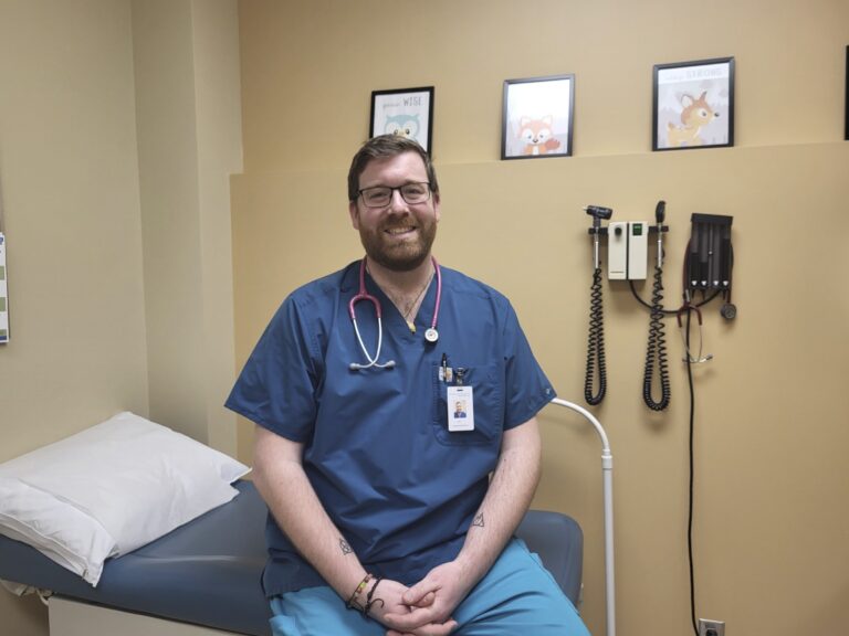 A man wearing blue scrubs sits on a patient bed and smiles.