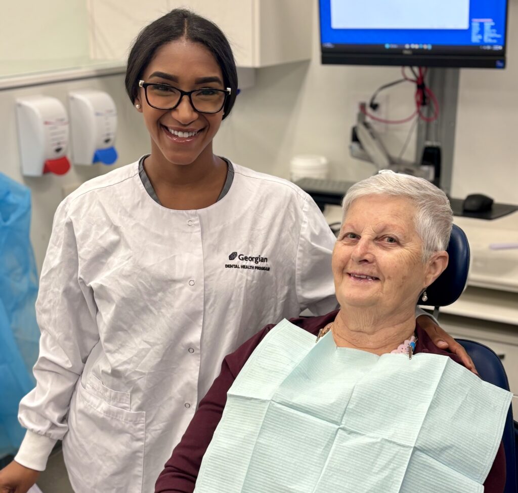 A person wearing a white lab coat reading "Georgian Dental Health Program" smiles next to a person sitting in a dental chair and wearing a blue paper bib. 