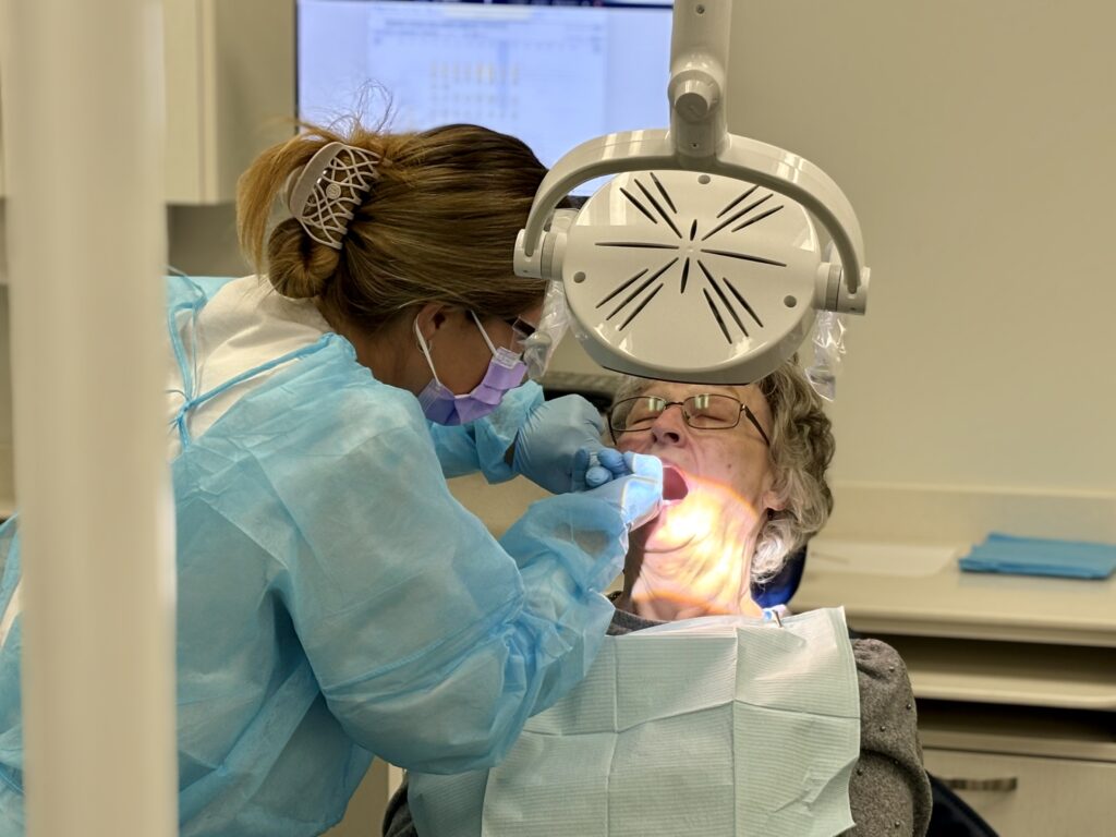 A person sits in a dental chair and has their mouth open as a person wearing protective wear works in their mouth. 