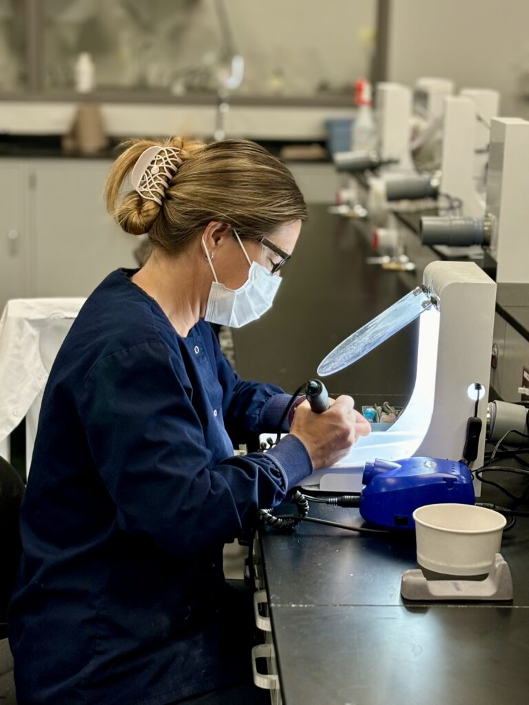 Side view of a person sitting at a long table, holding an electric tool and working on something underneath a screen. 