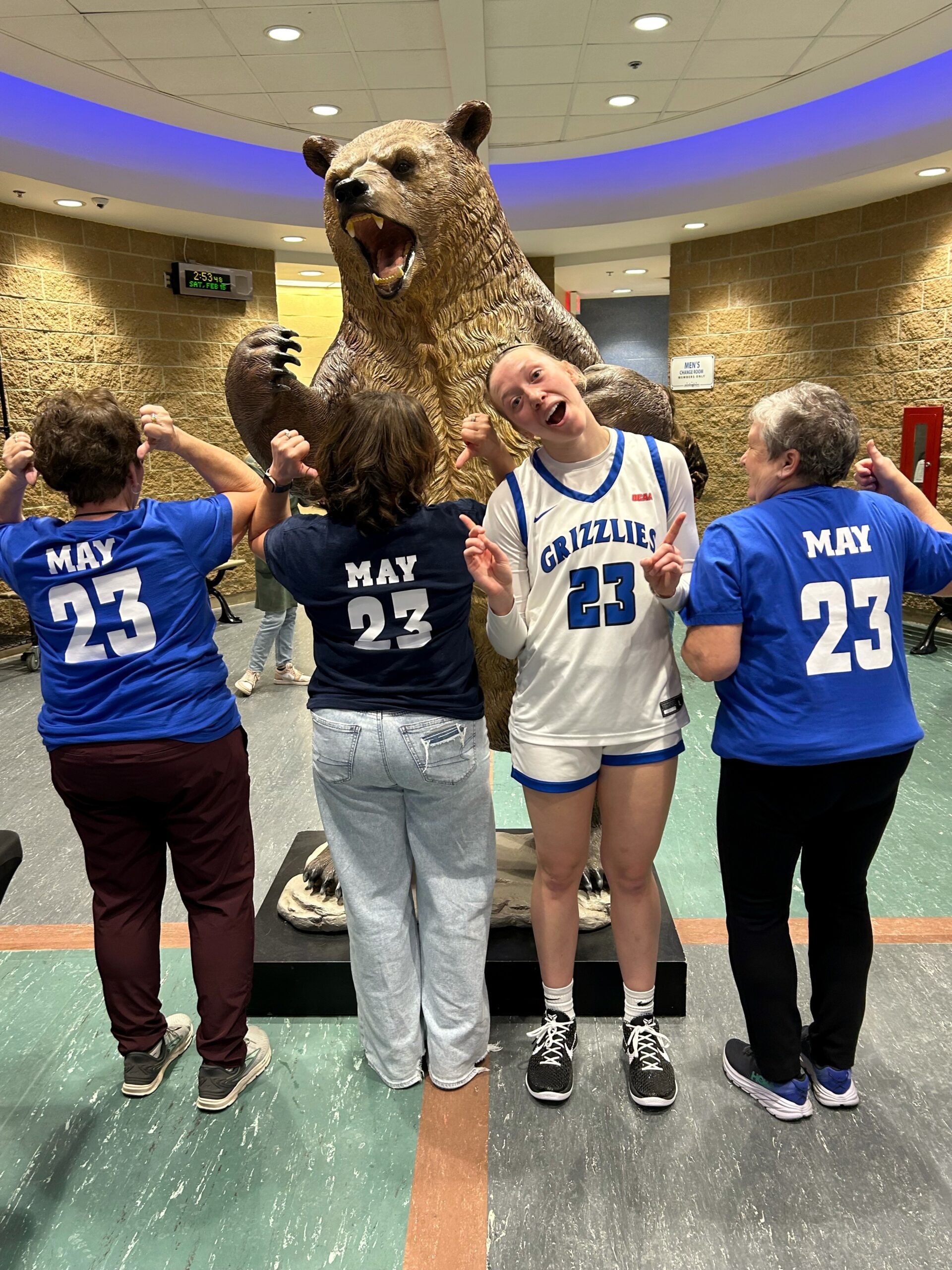 Four people stand together in front of a grizzly bear statue. One faces the camera wearing a basketball uniform reading "Grizzlies." The other three are facing away and the backs of their shirts read "May 23."