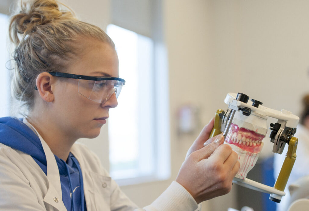 A person wearing a white lab coat and protective glasses works on a set of false teeth.