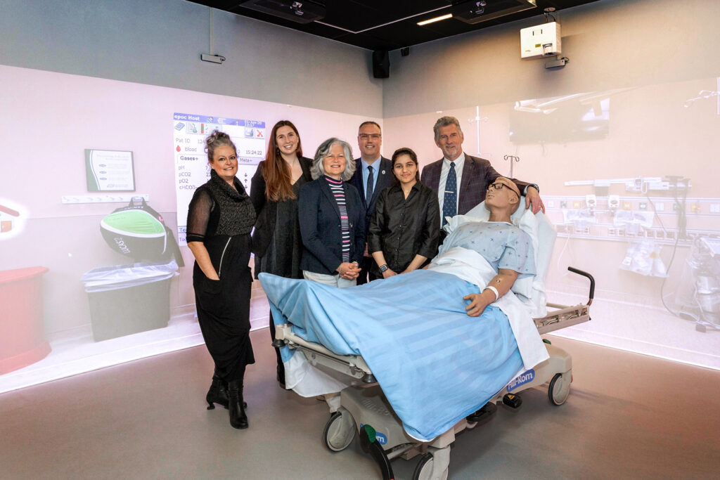 Six people stand together next to a hospital bed with a patient simulator lying in it, inside a room with projections of the inside of a hospital room on the walls. 