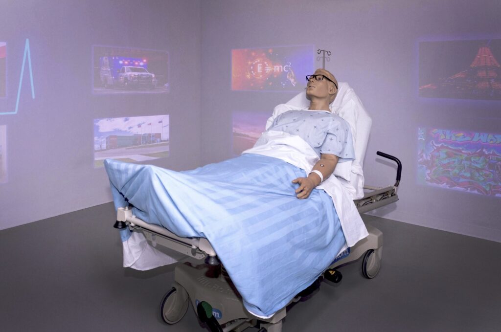 A simulator patient lies in a hospital bed in a simulation room with images projected onto the walls. 