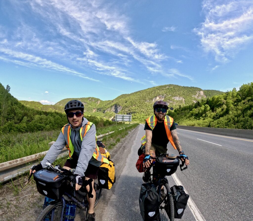 Two people ride bikes along a highway with treed hills in the background. 