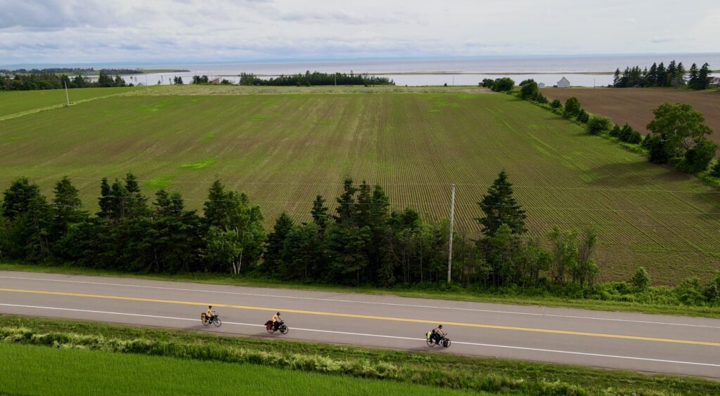 An aerial view of three people biking down a road next to green fields with a large body of water in the distance. 