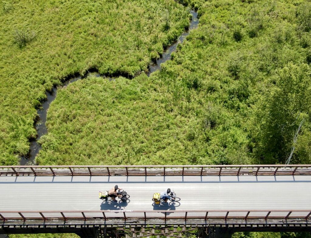 An aerial view of two cyclists riding on a bridge over green wetlands. 